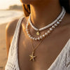 A close-up of a woman wearing a layered necklace featuring white shell beads and a gold starfish pendant.