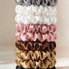 A stack of five satin hair scrunchies in various shades including gray, white, pink, brown, and black.