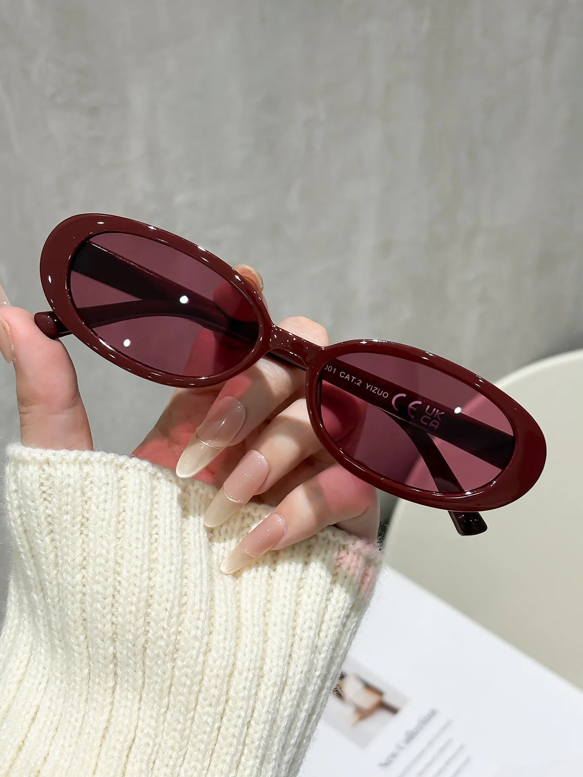 A hand holds a pair of maroon cat eye sunglasses with dark frames against a light background.