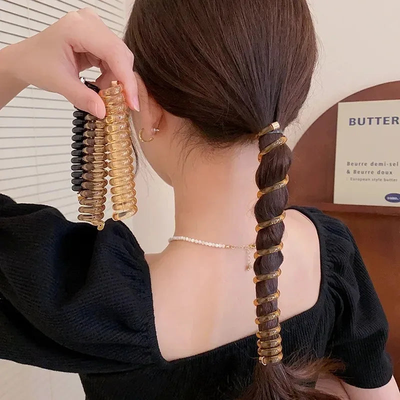 A hand is shown braiding a long dark hair extension into multiple gold rubber braided ponytail holders and scrunchies.