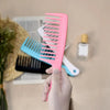 A hand holds a bright pink wide tooth detangling hair comb, with other combs and a bottle visible in the background.