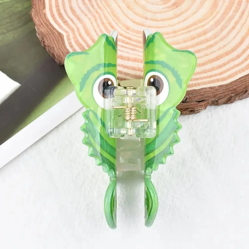 A green lizard-shaped claw clip hair accessory with large eyes and textured scales resting on a wooden surface.