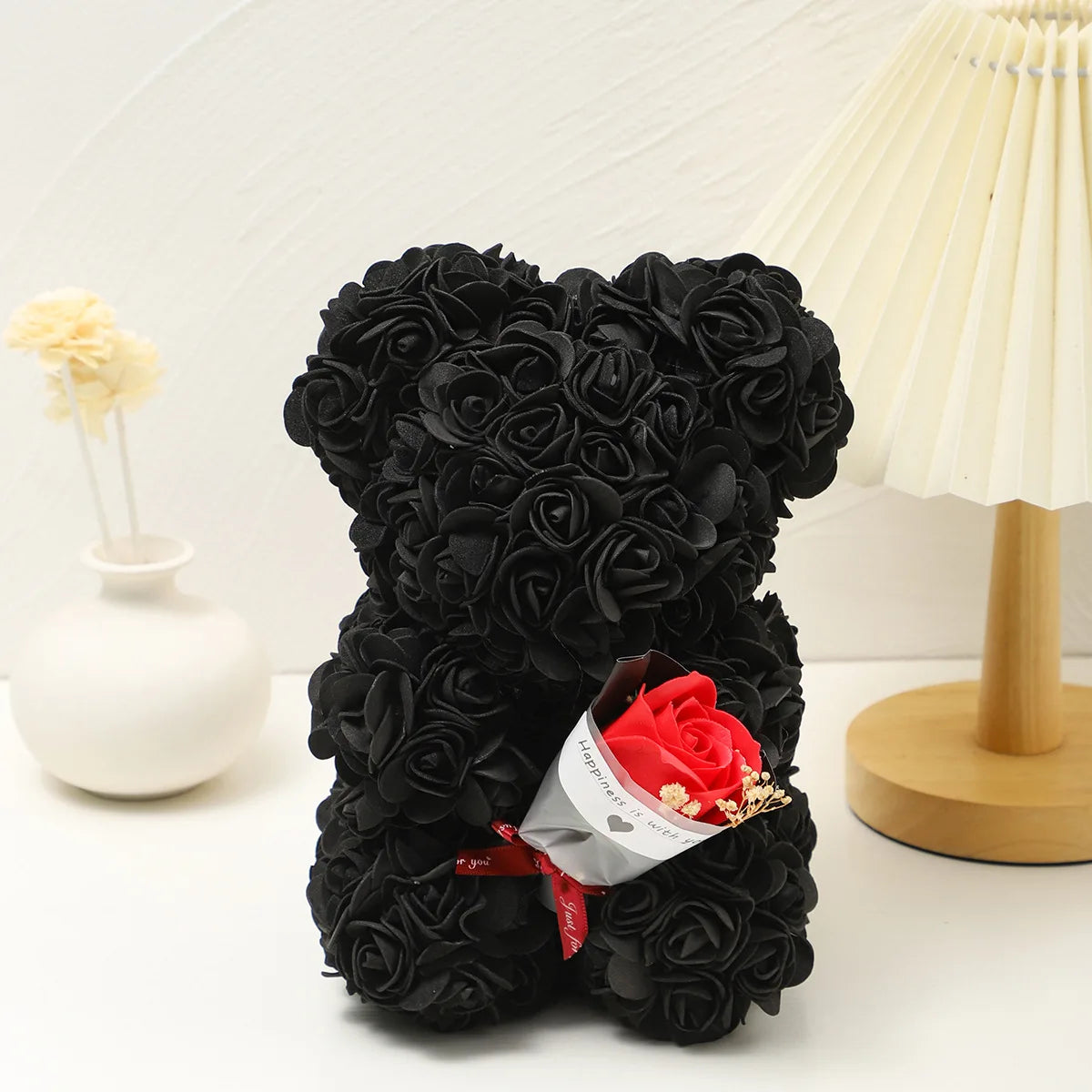 Un ours en peluche rose en mousse douce noir, à côté d'un vase blanc et d'une lampe de couleur crème.