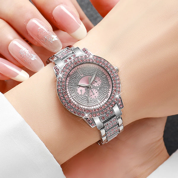 A silver watch with a pink dial and rose gold accents is worn on a wrist, displayed next to manicured hands.