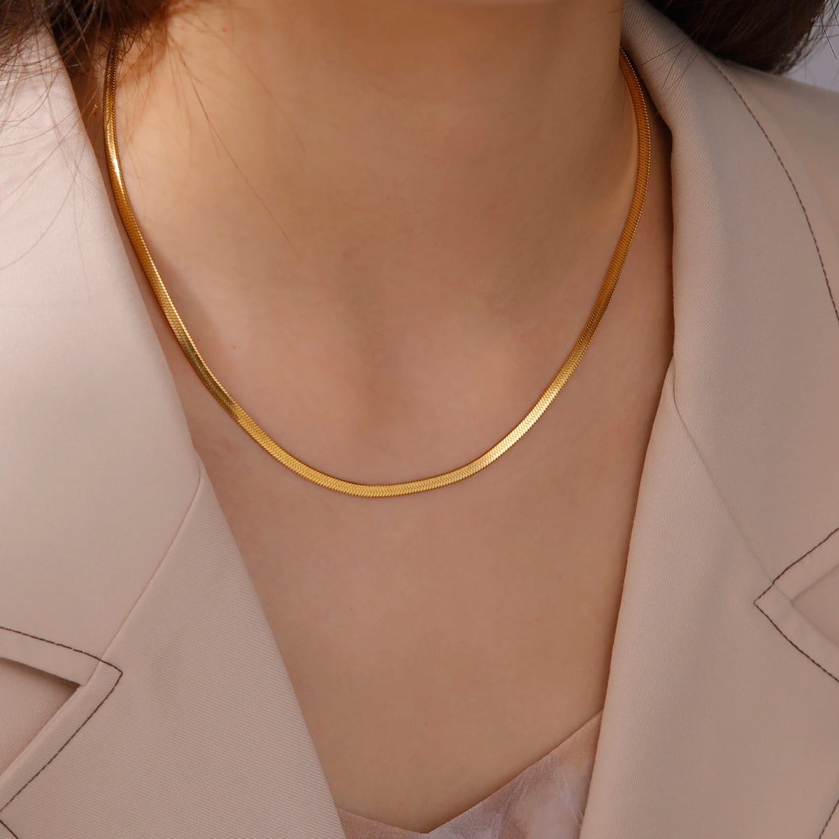 Nahaufnahme einer goldfarbenen Edelstahl-Schlangenkettenhalskette, kombiniert mit einem hellbeigen Blazer.