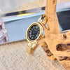 A gold-toned quartz watch with a black face and blue hands is displayed next to a piece of natural wood.