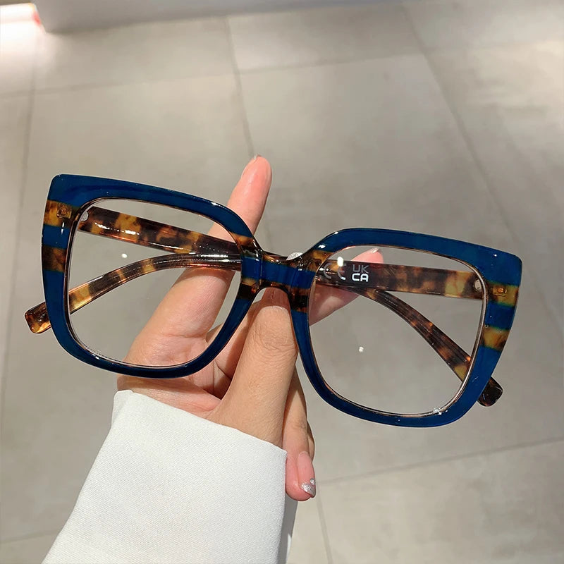 Eine Hand hält eine modische, große, quadratische Brille mit Schildpatt- und blauen Akzenten.