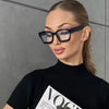 Una mujer con gafas cuadradas negras contra un fondo gris.