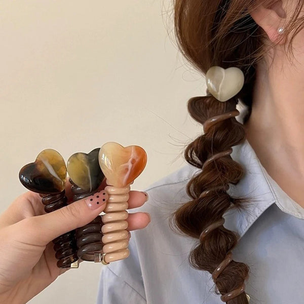 Una mano sostiene un accesorio para el cabello trenzado en forma de corazón, hecho de piedra natural en tonos marrones y ámba
