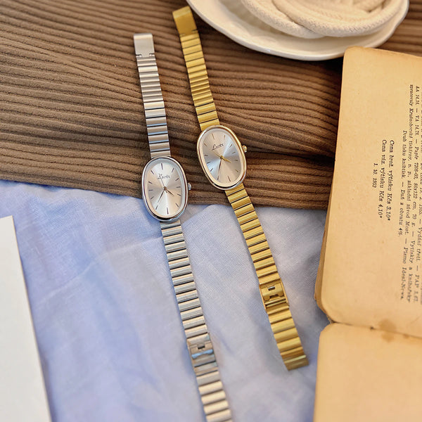 A silver-toned quartz watch with a gold bracelet is displayed next to an open book on a light blue cloth.
