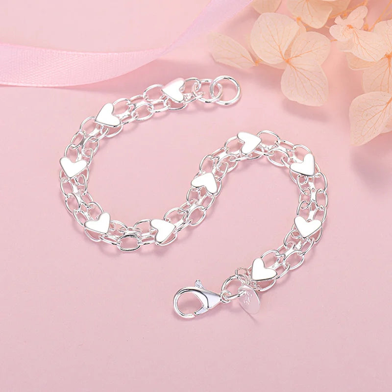 A silver heart charm bracelet made of sterling silver links and charms is displayed on a pink background.