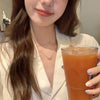 A woman wearing a white shirt is holding an orange drink and displaying a pink crystal heart necklace.