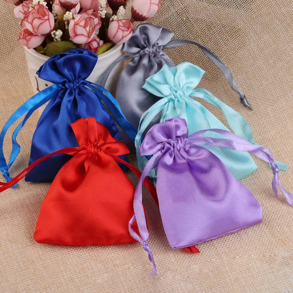 A collection of satin drawstring jewelry pouches in blue, gray, and pink are displayed on a beige fabric background.