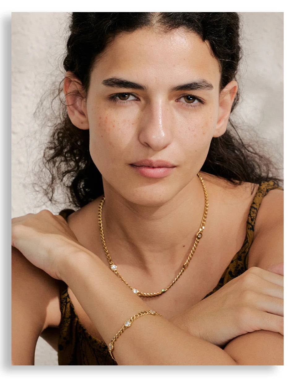 A close-up portrait of a woman wearing a gold stainless steel charm necklace.