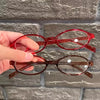 Une main tient des lunettes de mode ovales rétro teintées en rouge sur un fond de briques grises.
