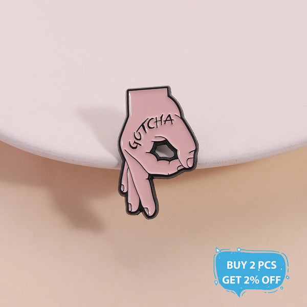 A pink enamel lapel pin shaped like a hand making the 'gotcha' gesture.