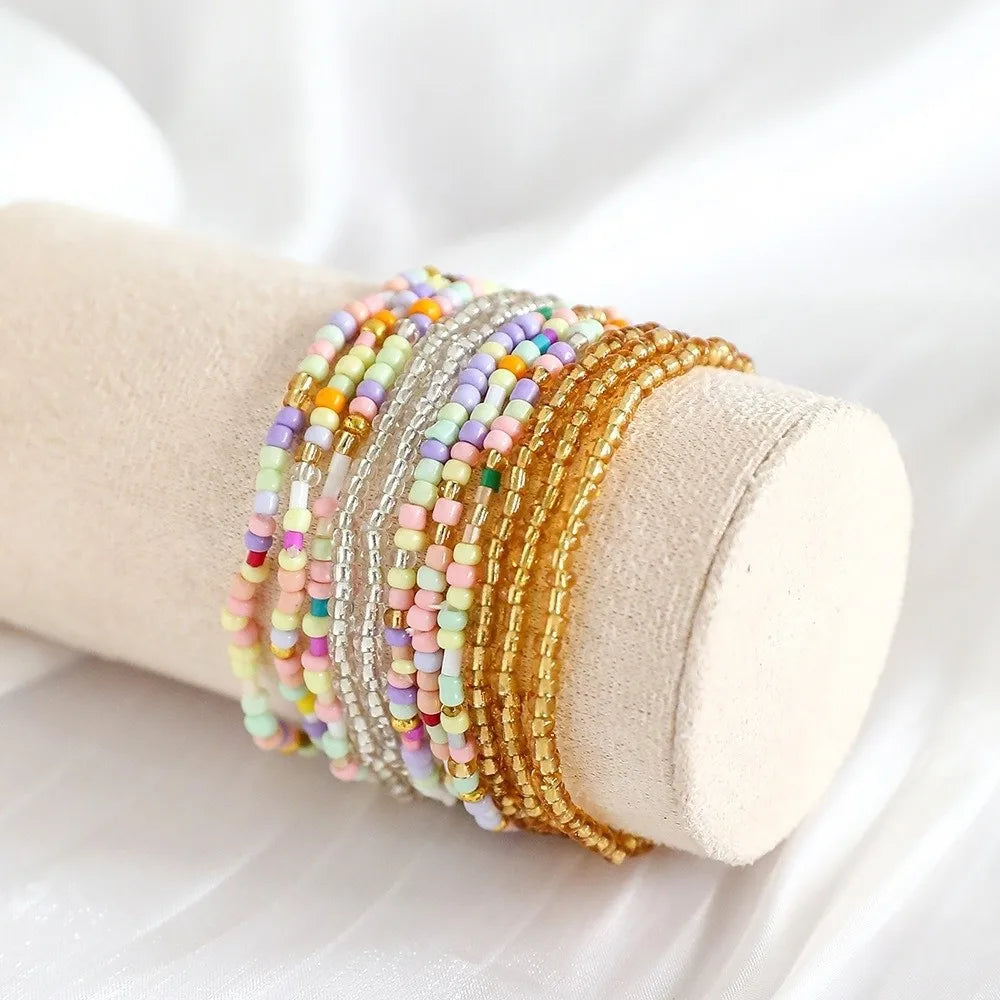 A stack of colorful beaded bangles, including pink, yellow, and gold tones, is displayed on a beige cylindrical object.
