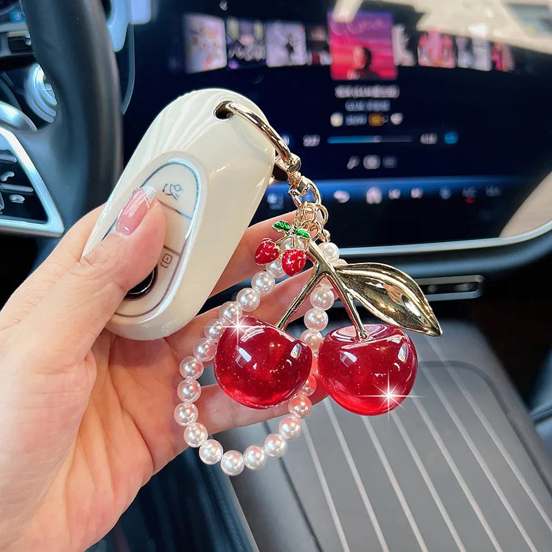 A hand holds a white key chain decoration featuring red cherry beads and pearl strands.