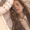 A model is wearing several delicate gold-plated foot chains adorned with small blue and purple crushed stones.