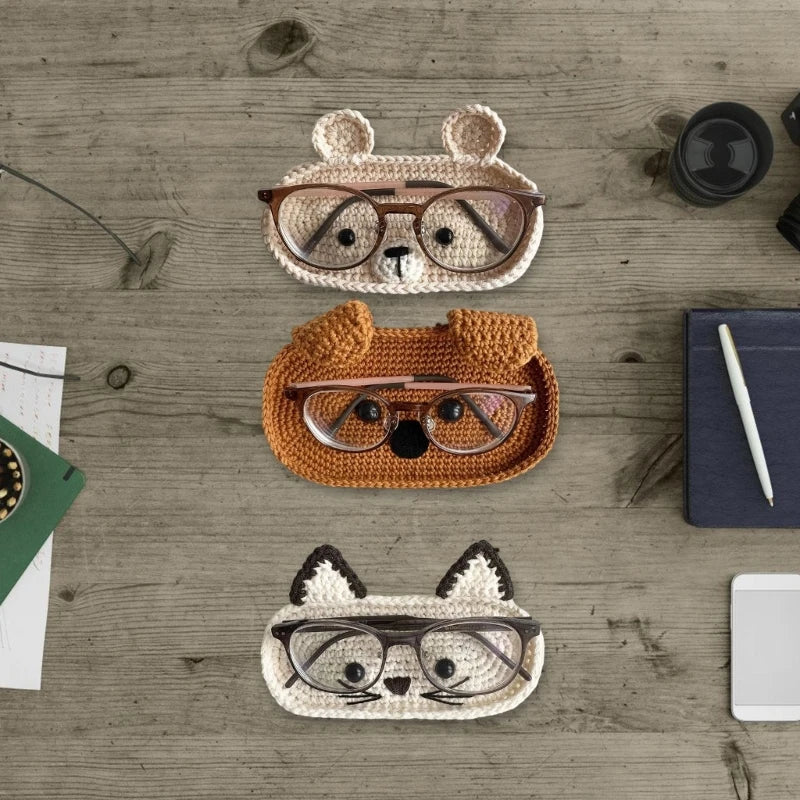 Three handmade crochet animal eyeglass storage cases in various colors and styles resting on a wooden surface.