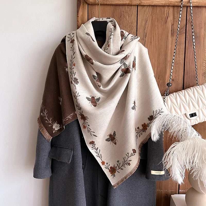 A beige pashmina shawl with a floral print is draped over a gray coat, displayed against a wooden background.