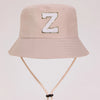 A light beige sun hat with a large gold letter Z embroidered on the front and a light beige strap.