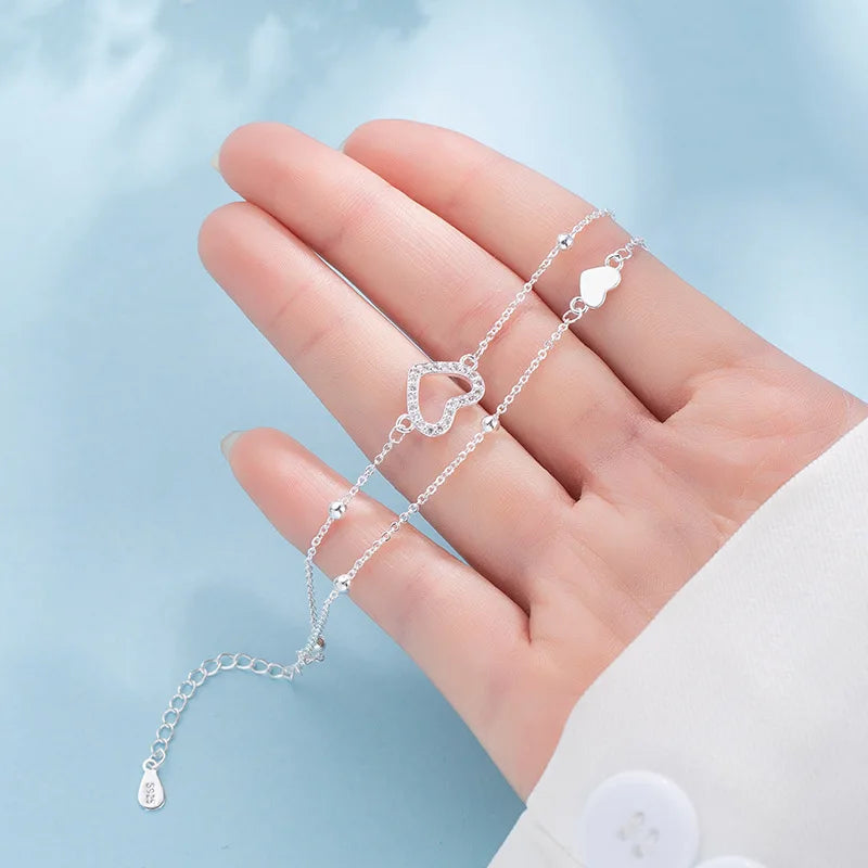 A hand is holding a sterling silver double heart charm bracelet with a delicate chain and charms against a light blue backgro