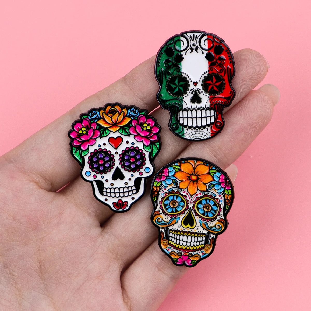 Three colorful enamel skull pins featuring vibrant flowers and detailed designs on a hand.