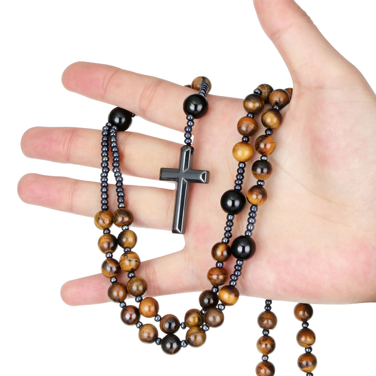 A hand holds a rosary necklace featuring tiger eye and black agate beads with a hematite cross pendant.