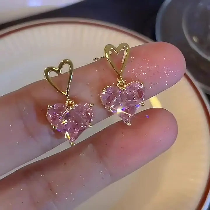 A hand holds a pair of gold-toned heart-shaped necklaces featuring pink crystal stones.
