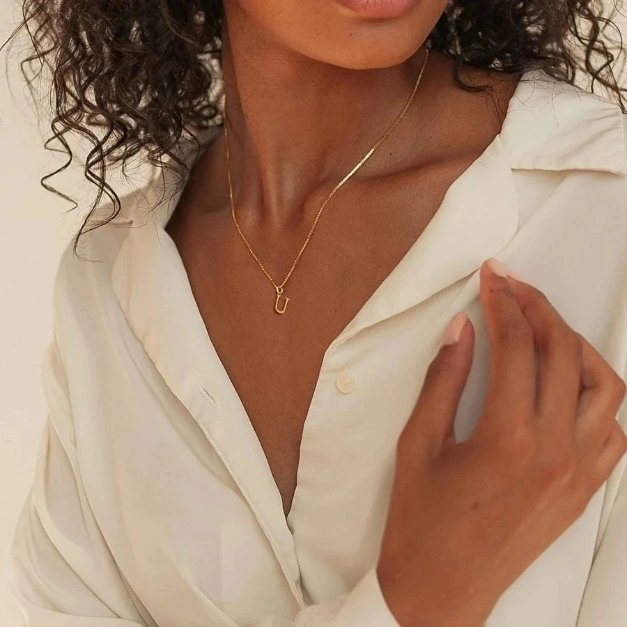 A close-up of a woman wearing a delicate gold initial letter necklace with a small charm against a light colored shirt.