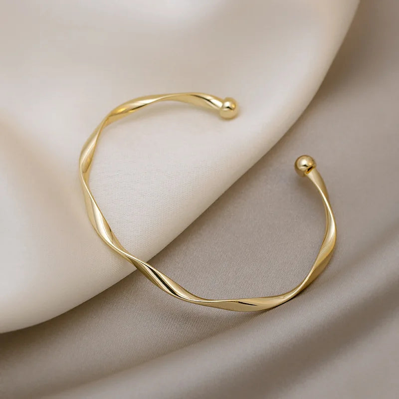 A minimalist gold-tone twisted cuff bangle resting on a neutral beige fabric.
