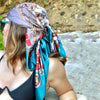 A woman wearing a teal silk print shawl draped around her head and neck, outdoors.