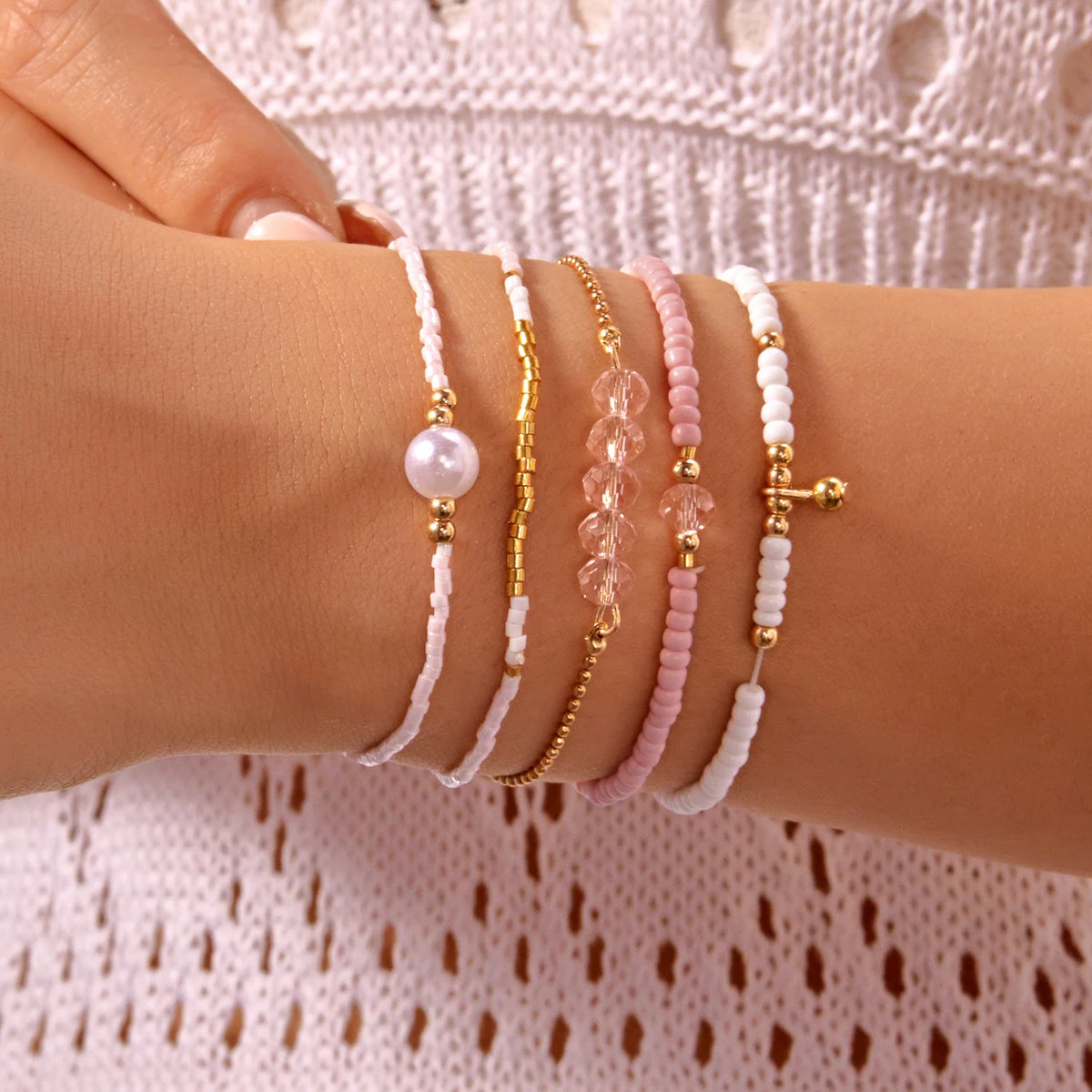 A hand is wearing a stack of delicate pearl and pink beaded bracelets on a wrist.