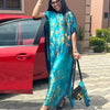 Eine Frau in einem tealfarbenen Seidenkaftan mit goldener Blumenmuster steht neben einem roten Auto.