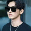 A close-up portrait of a young man wearing black square frame sunglasses against a blurred background.
