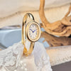 A gold-toned quartz watch with a round face and Roman numerals is displayed on white lace fabric.