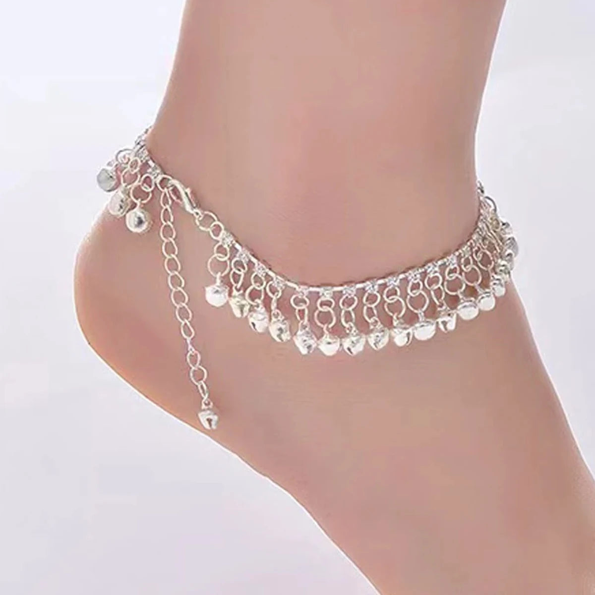 A delicate silver bell foot chain anklet worn around a slender ankle.