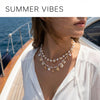 A close-up of a woman wearing a layered boho necklace featuring starfish and shell charms against a bright outdoor background
