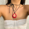 A woman wearing a black rope choker necklace with a red pendant against a light background.