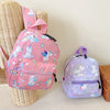 A pink and purple unicorn dinosaur pattern kids backpack is displayed next to a woven basket.