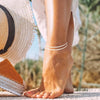 A close-up of a person's foot wearing delicate silver beaded anklets, standing outdoors.