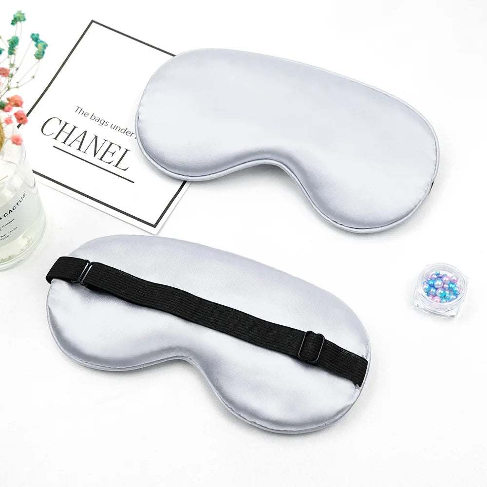 A pair of silver silk sleep masks with black straps are displayed next to a box labeled Chanel.