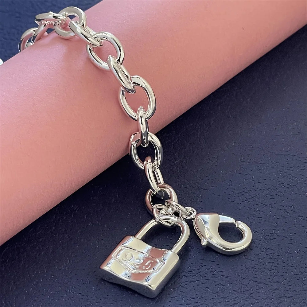 A silver charm lock bracelet with a chain and padlock is displayed on a dark surface next to a pink cylindrical object.