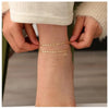 A close-up of a woman's ankle showing layered gold letter anklets being worn.