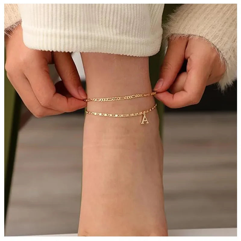 A close-up of a woman's ankle showing layered gold letter anklets being worn.