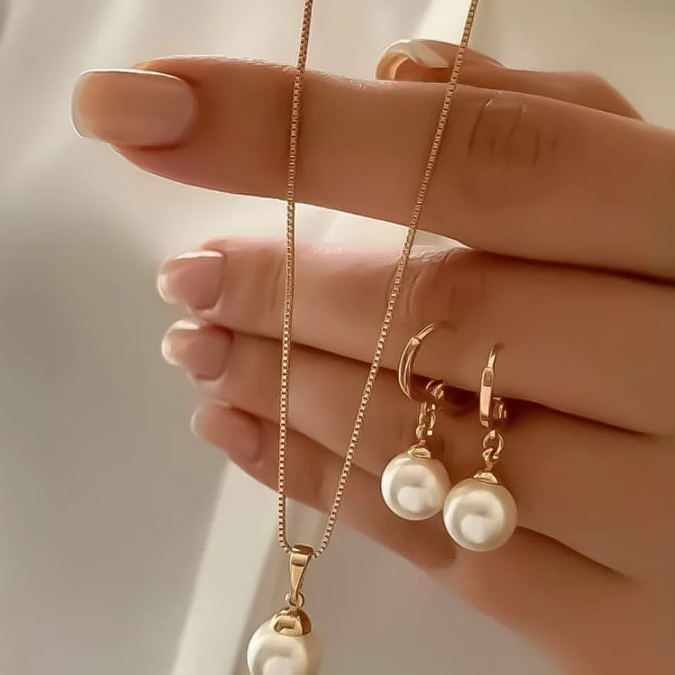 A close-up shot of a hand wearing a dainty gold pearl jewelry set featuring matching earrings and a delicate necklace.