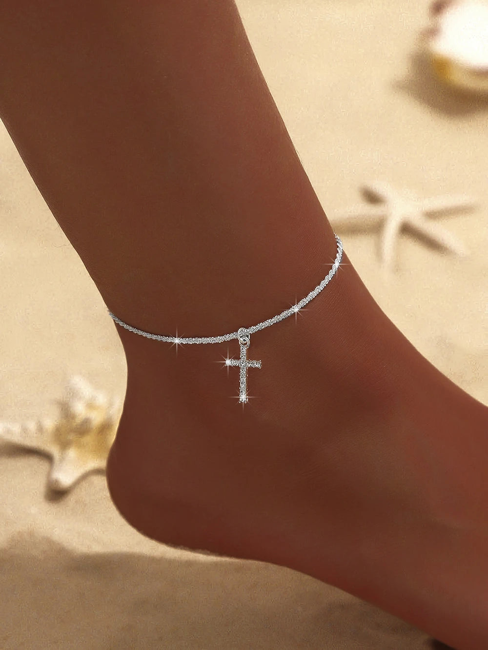 A close-up of a sparkling zircon heart cross anklet worn on a person's ankle against a light background.