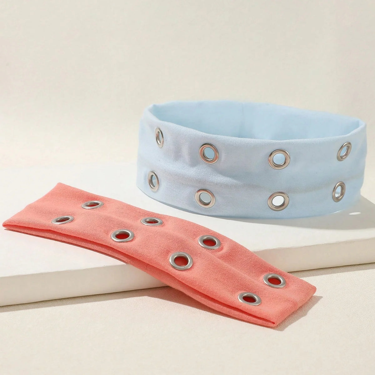 A light blue metal hollow headband and a coral pink fabric headband are displayed on a white surface.