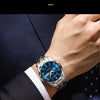 A close-up of a luxury men's quartz watch with a blue luminous dial and silver bracelet worn on a wrist, next to a dark suit 